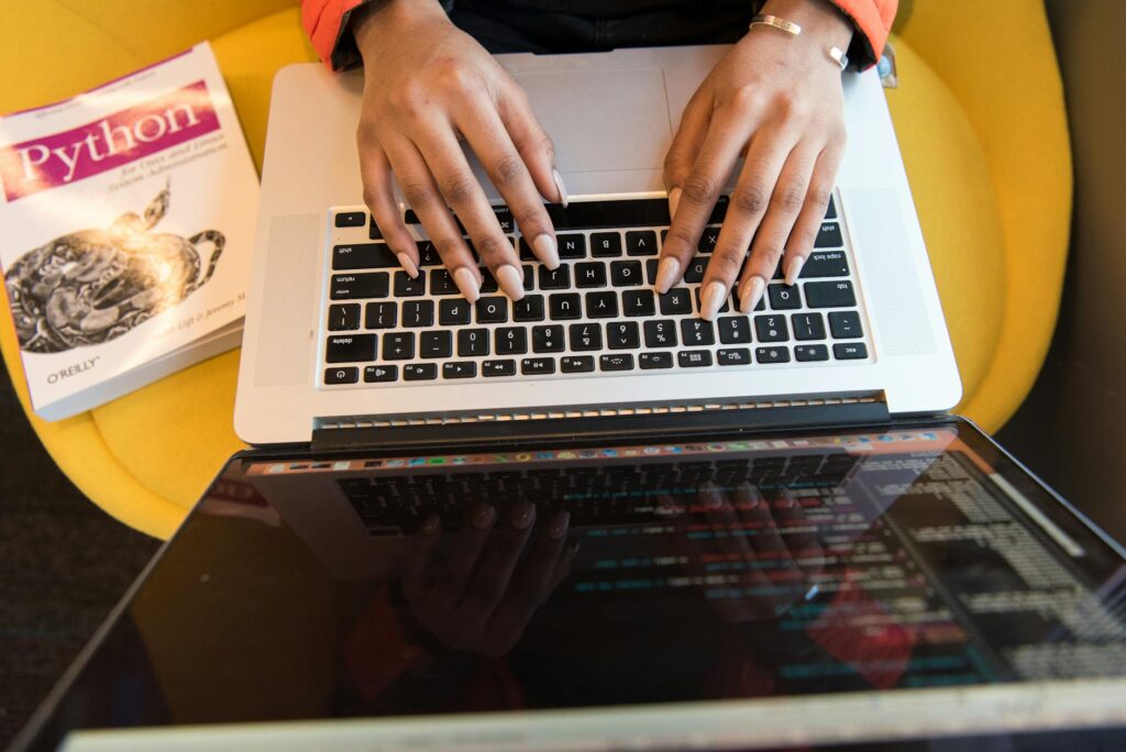 Close-up of hands typing on a laptop keyboard, Python book in sight, coding in progress. (Photo by Christina Morillo on Pexels)