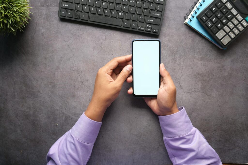Man in purple shirt holding a smartphone with blank screen at office desk with keyboard, notebook... (Photo by Towfiqu barbhuiya on Pexels)