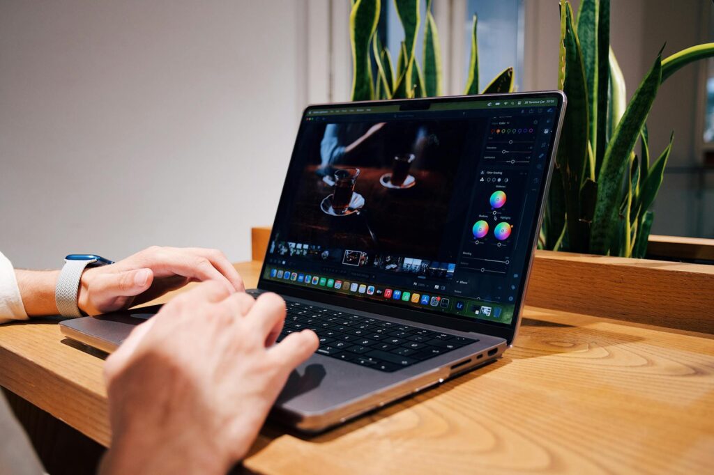 Close-up of hands editing a photo on a laptop in an office setting, surrounded by green plants. (Photo by iam hogir on Pexels)