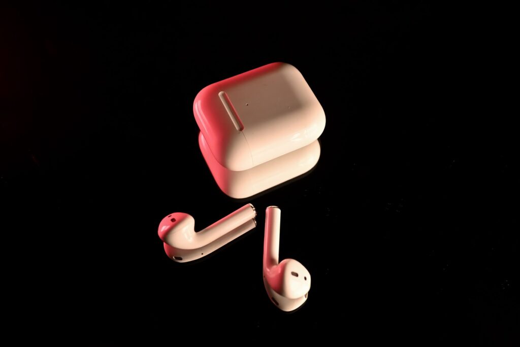 White wireless earbuds with a charging case, highlighted with red lighting on a reflective black ... (Photo by HS Studio By Hussnain on Pexels)