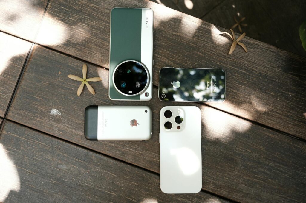 Assorted smartphones displayed outdoors on a wooden table under dappled sunlight. (Photo by Phong Thanh on Pexels)