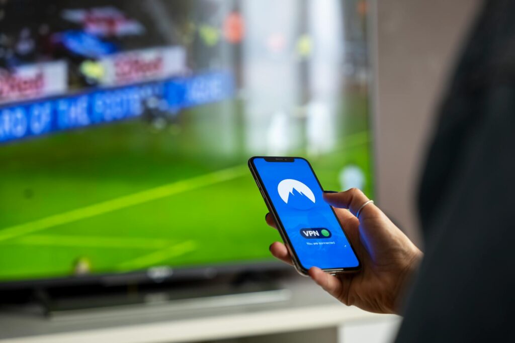 Close-up of a person holding a smartphone with a VPN app, streaming sports on TV. (Photo by Stefan Coders on Pexels)