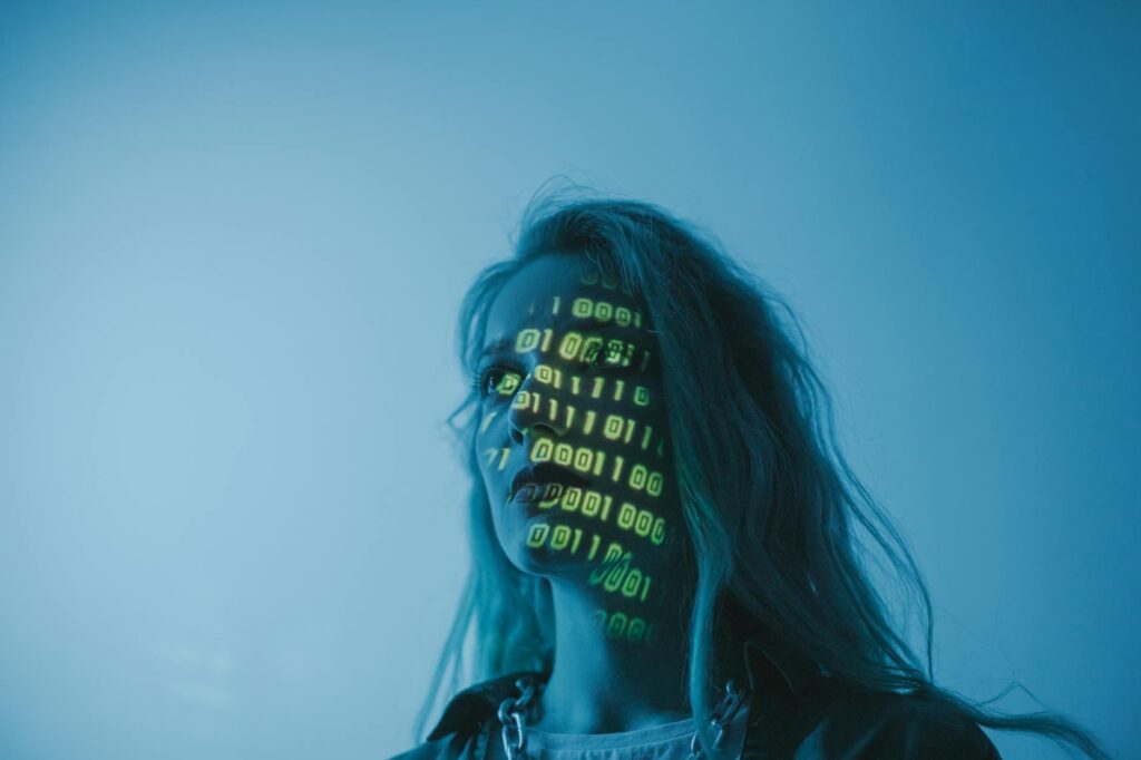 A woman with binary code lights projected on her face, symbolizing technology. (Photo by cottonbro studio on Pexels)