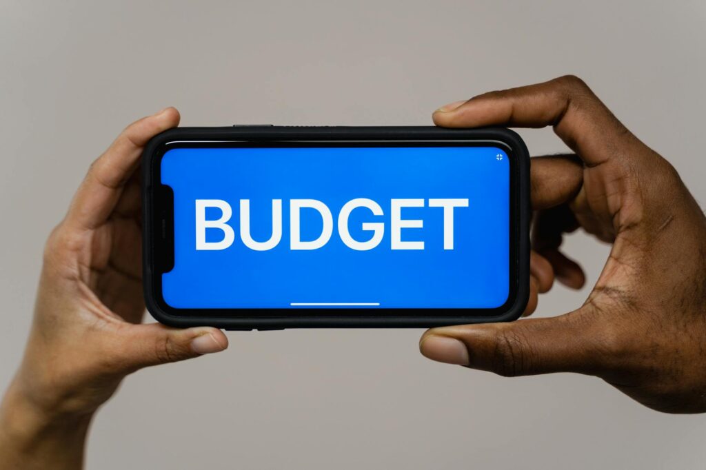 Two hands hold a smartphone displaying the word 'budget' on a blue screen, symbolizing financial ... (Photo by Tima Miroshnichenko on Pexels)