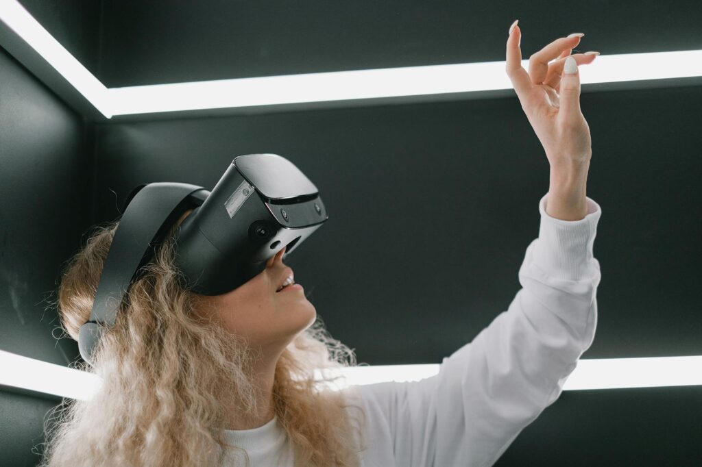 A woman wearing a headset engages with virtual reality, reaching out her hand. (Photo by SHVETS production on Pexels)