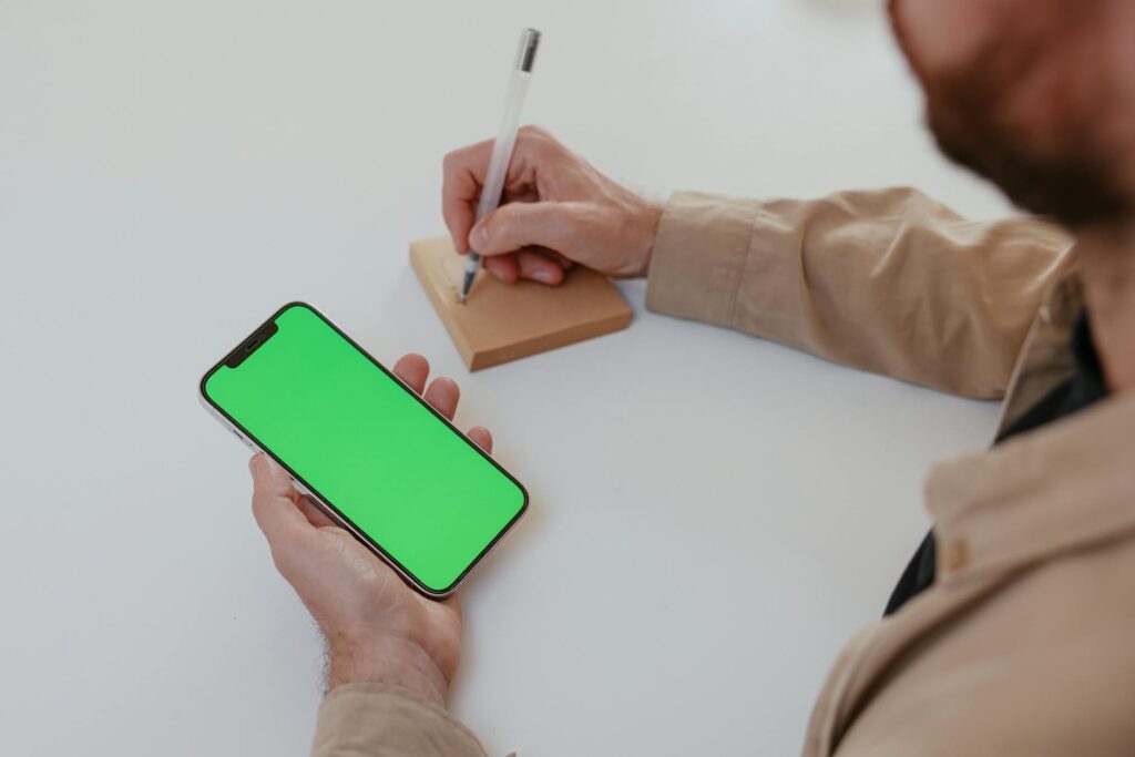 A person writing on a notepad while holding a smartphone with a green screen, ideal for mockups. (Photo by Cup of  Couple on Pexels)