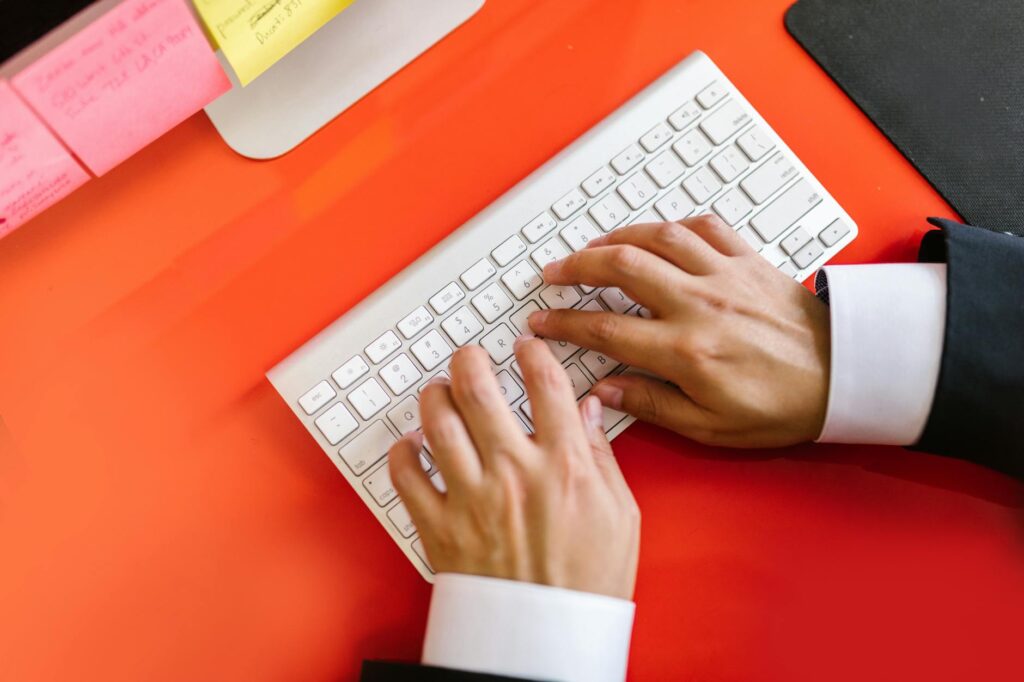 Close-up of hands typing on a wireless keyboard on an orange desk, ideal for tech and business th... (Photo by RDNE Stock project on Pexels)