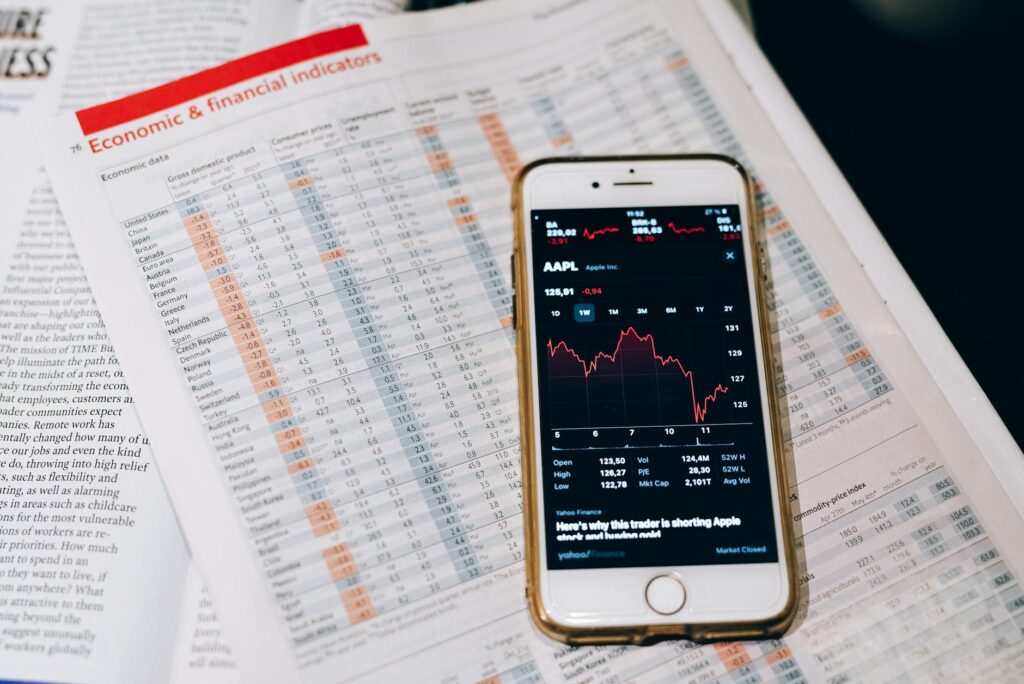 Smartphone displaying stock market data on papers with financial charts. (Photo by Leeloo The First on Pexels)