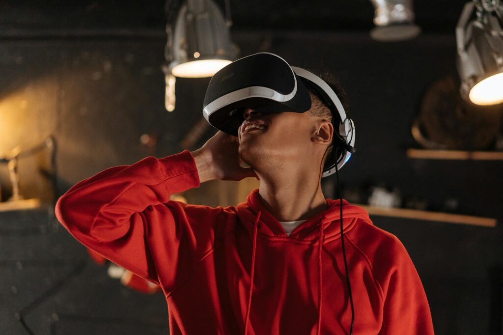 A young man wearing a red hoodie explores virtual reality indoors with a VR headset. (Photo by Mikhail Nilov on Pexels)