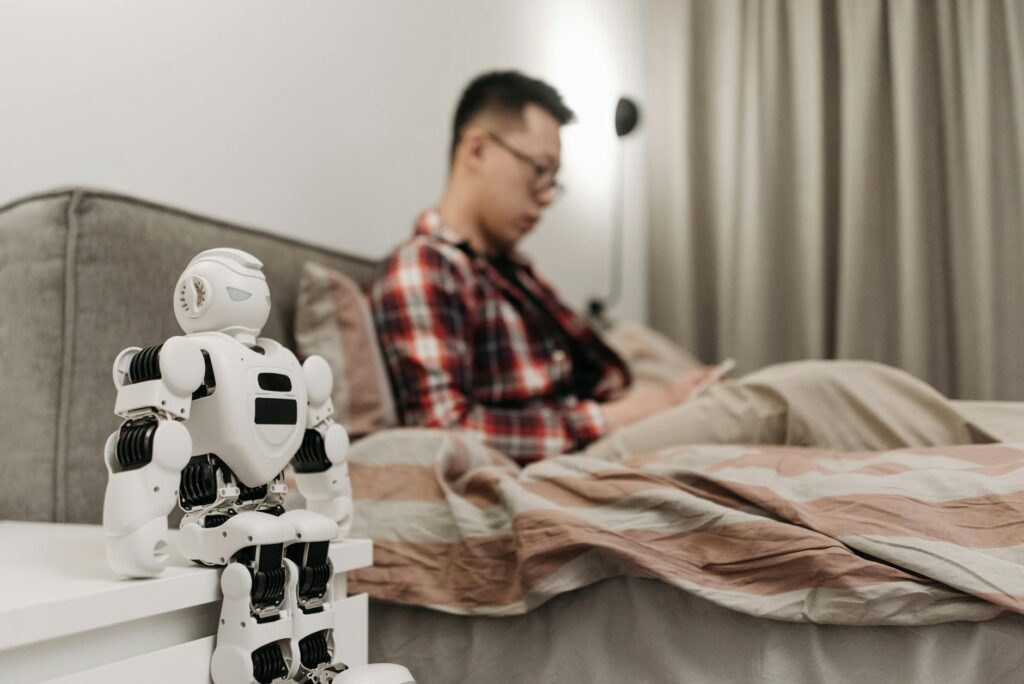 A modern bedroom featuring a robot on a bedside table and a man sitting on a bed. (Photo by Pavel Danilyuk on Pexels)